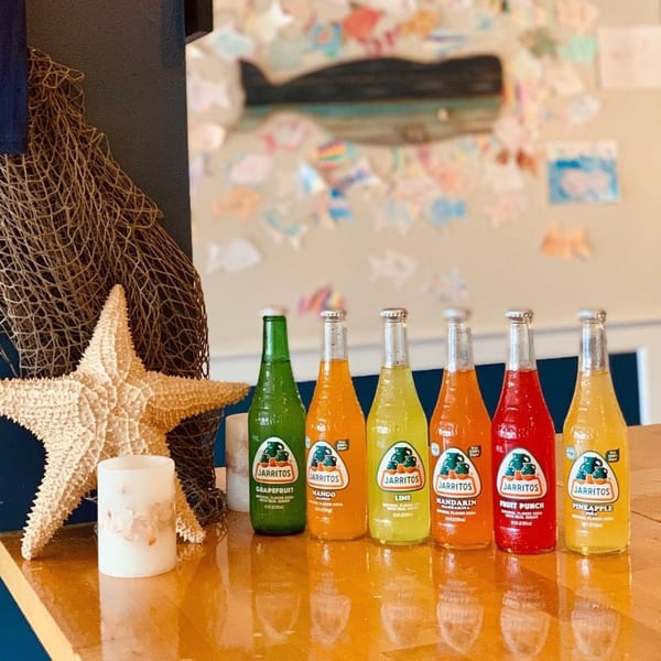 Six colorful Jarritos sodas on a wooden surface with a starfish and fishing net.
